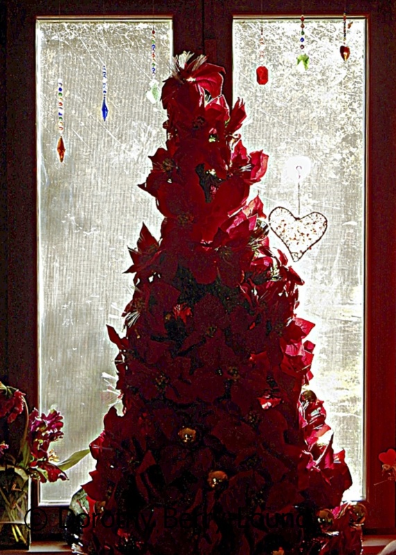 A red artificial christmas tree with lights and baubles, sits in a window.