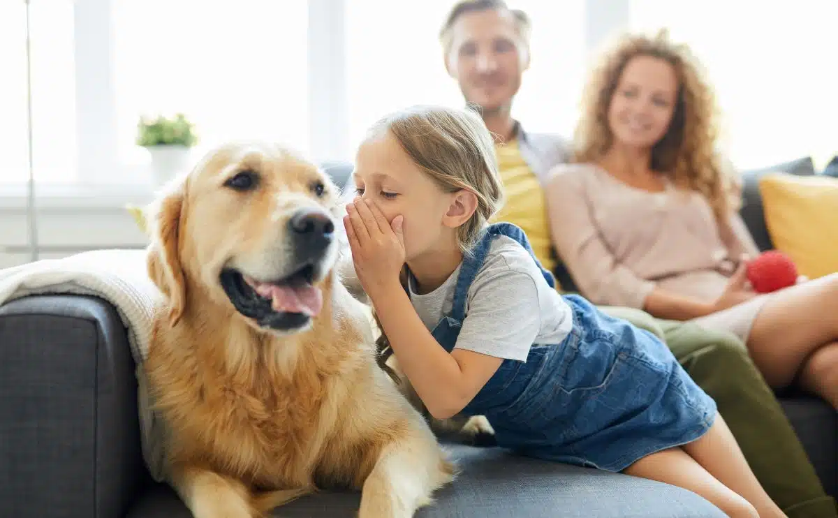 girl whispering into a goldens ear with family behind her all sitting on a sofa